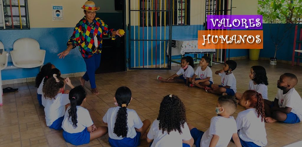 Crianças assistindo professora vestida com chapéu e roupa colorida dançando.